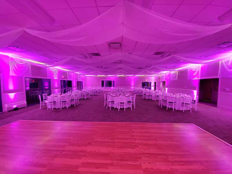 Dining room shown in all pink lighting.  3 rows of table seating 5, 4 table deep for a total of 12 tables.  Curtains drape ceiling and walls, dance floor is set up in front of tables